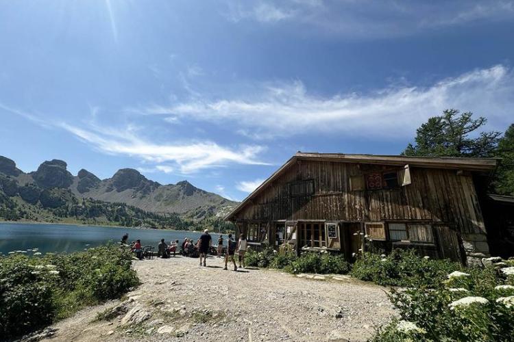 Vue de côté du refuge en bois, terrasse avec tables et chaises. Lac d'Allos visible à proximité, avec les Tours du lac en fond de photographie - Vue de côté du refuge en bois, terrasse avec tables et chaises. Lac d'Allos visible à proximité, avec les Tours du lac en fond de photographie